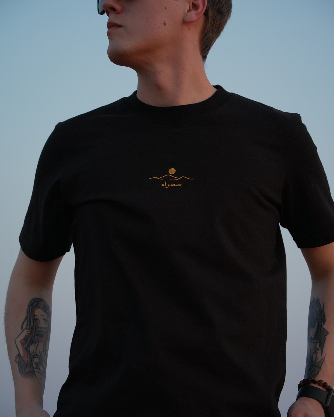 The Al Quaa Galaxy tee, front, worn in the dunes — the embroidered sun-and-dune mark on the chest