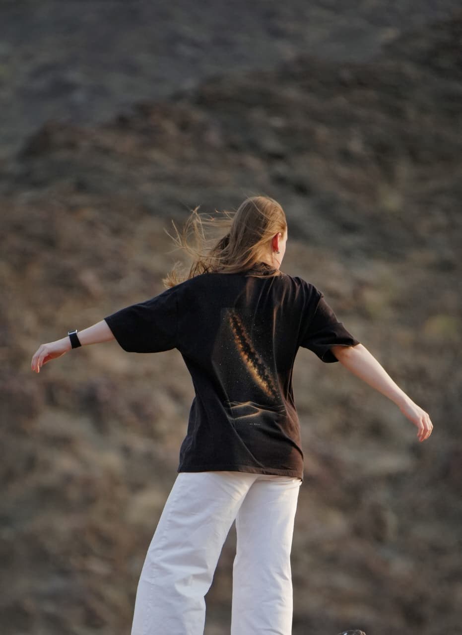 The Al Quaa Galaxy tee from behind, arms out against the mountains — the Milky Way print across the back. Female model wears M in the Oversized fit.