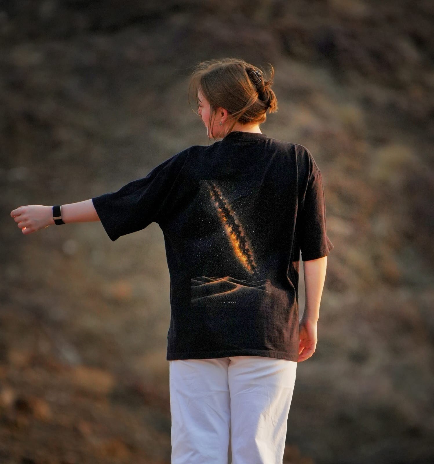 The back of the Al Quaa Galaxy tee in the mountains at dusk, the Milky Way print catching the last light. Female model wears M in the Oversized fit.