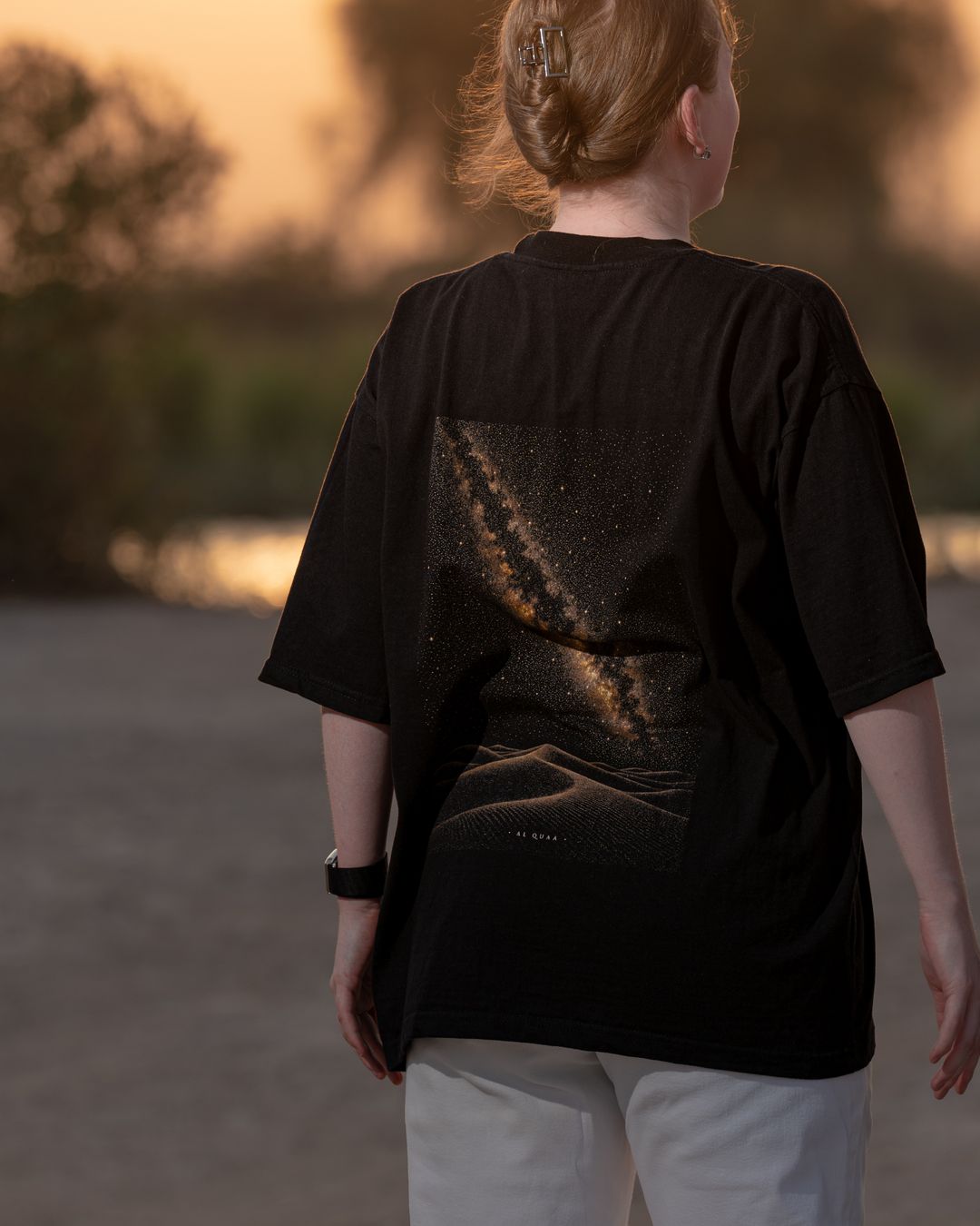 The Al Quaa Galaxy tee from behind by the lake — the Milky Way print across the back. Female model wears M in the Oversized fit.