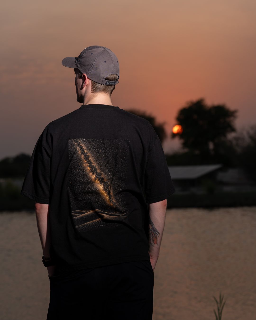 The Al Quaa Galaxy tee from behind as the sun sets over the lake — the Milky Way print across the back. Male model wears M in the Oversized fit.
