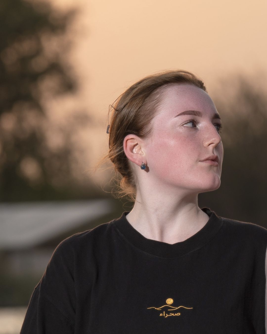 The Al Quaa Galaxy tee, front — the gold embroidered sun-over-dunes mark and صحراء on the chest. Female model wears M in the Oversized fit.