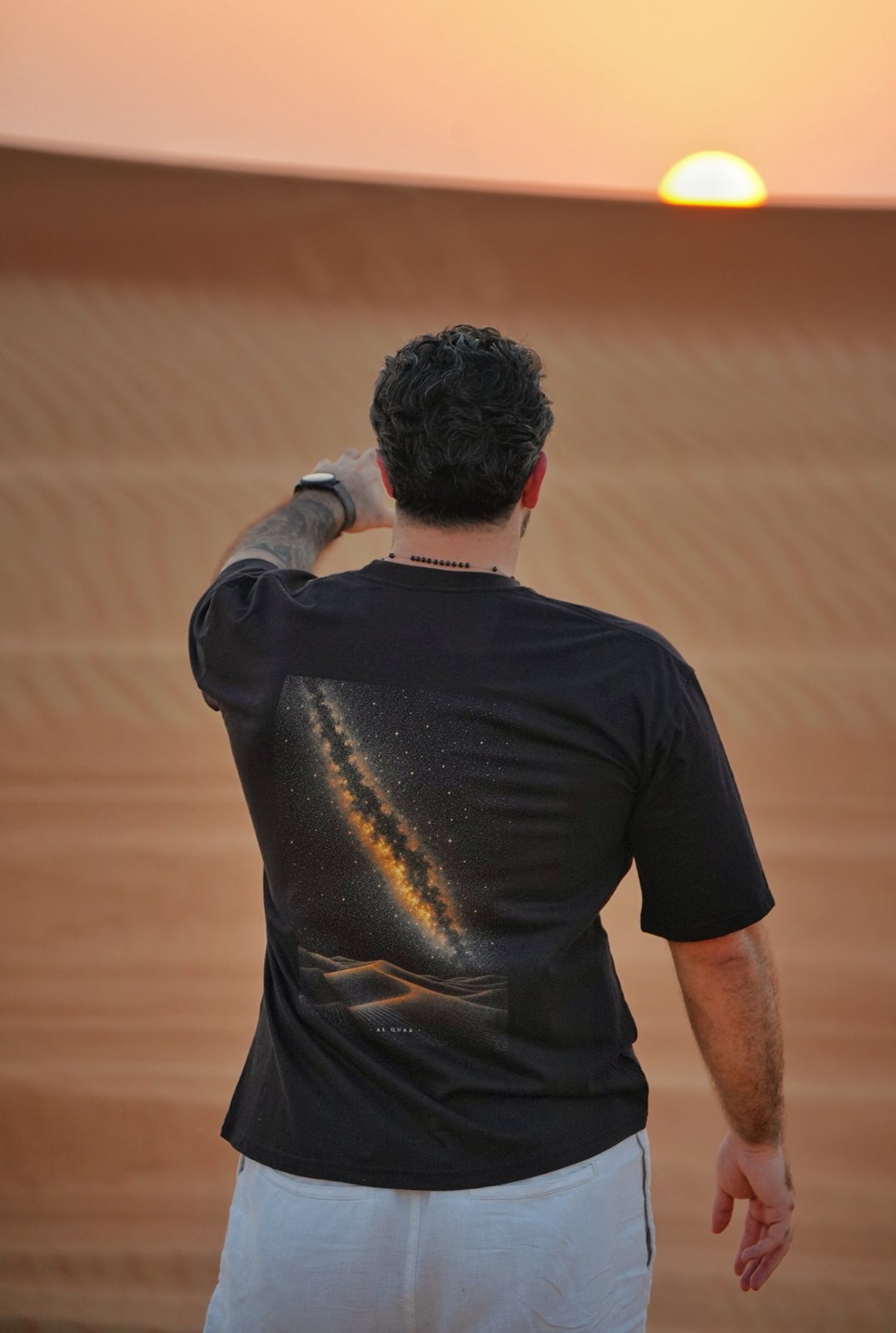 The Al Quaa Galaxy tee worn at sunset — the Milky Way print across the back as the sun drops behind the dunes