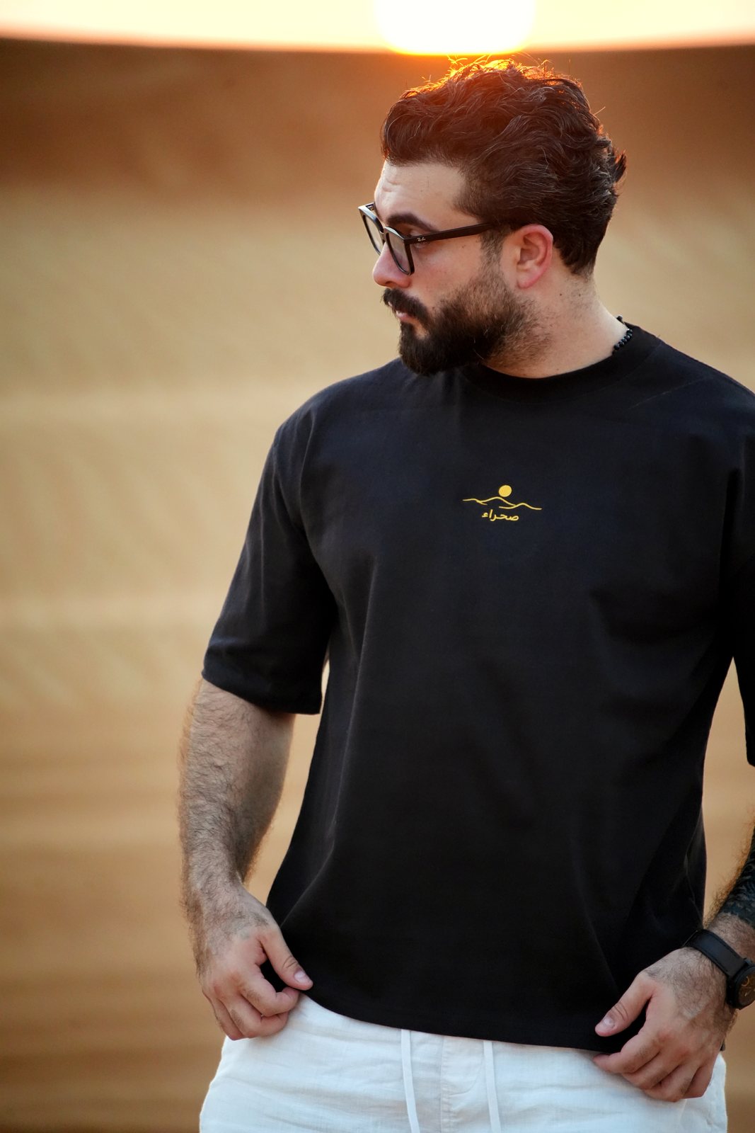 The Al Quaa Galaxy tee worn in the dunes — the gold embroidered sun-over-dunes mark and صحراء on the chest