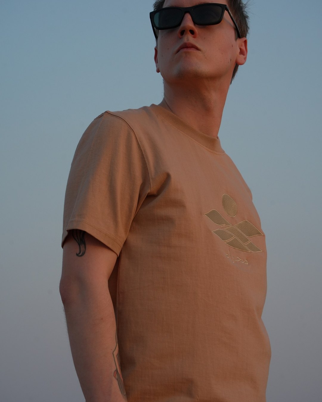 The Empty Quarter tee, chest close in the dunes — the tonal embroidery catching the low sun