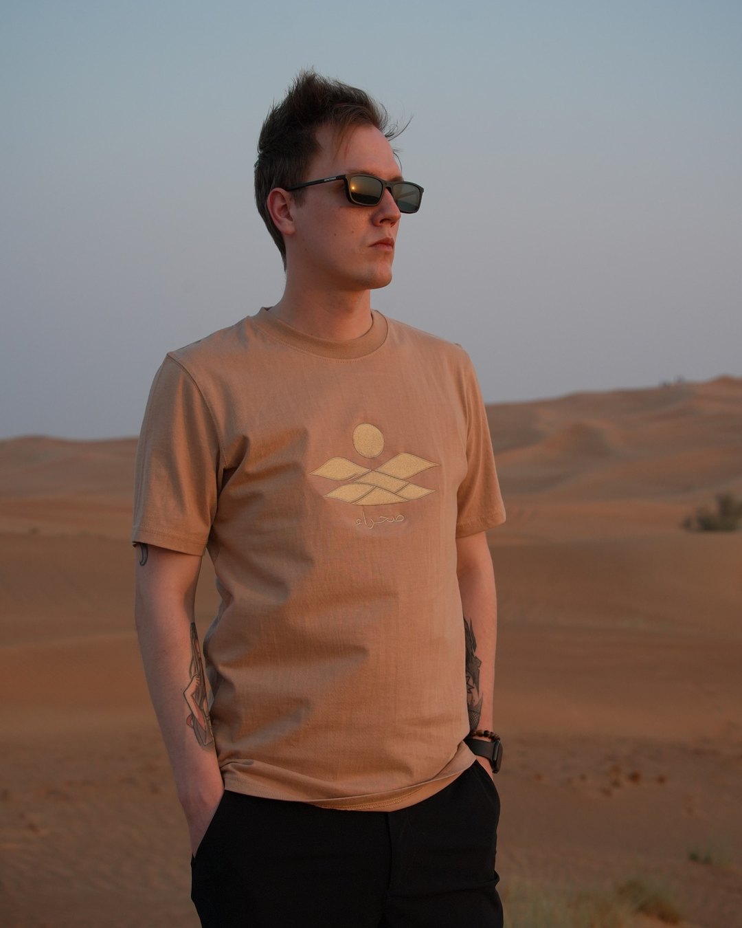The Empty Quarter tee worn in the dunes at sunset — the tonal embroidered dunes, sun and صحراء on the chest