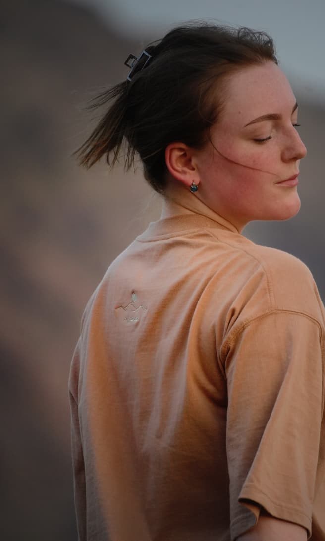 The Empty Quarter tee worn in the mountains, in profile — the tonal embroidered sun and dunes on the chest. Female model wears M in the Oversized fit.
