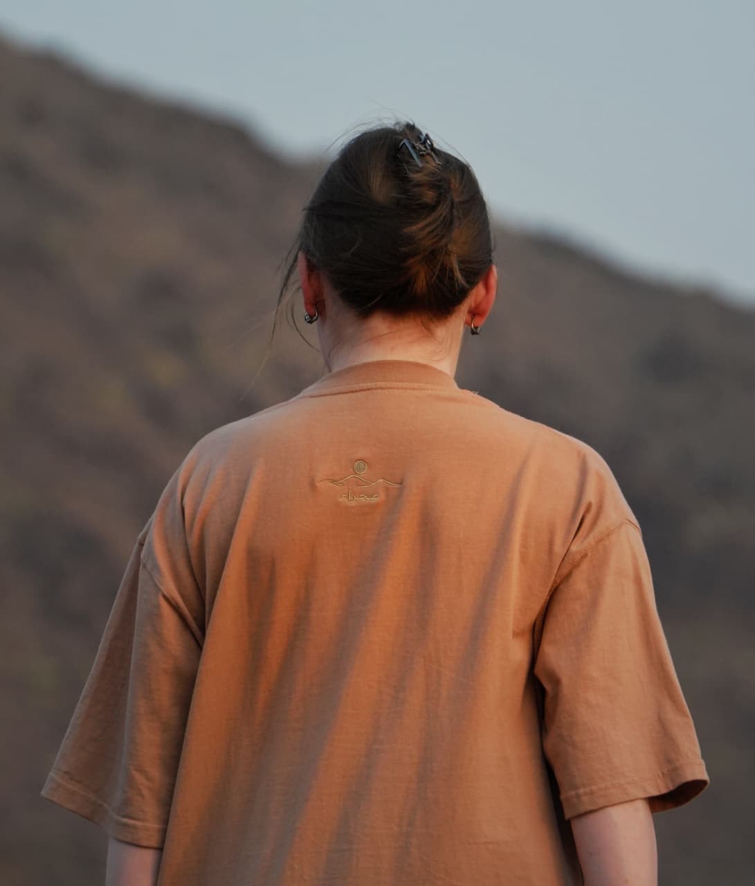 The back of the Empty Quarter tee in the mountains — plain, with the back-neck embroidery. Female model wears M in the Oversized fit.