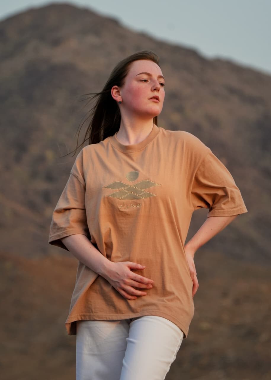 The Empty Quarter tee worn on a mountain ridge — the tonal embroidered sun over the dunes across the chest. Female model wears M in the Oversized fit.