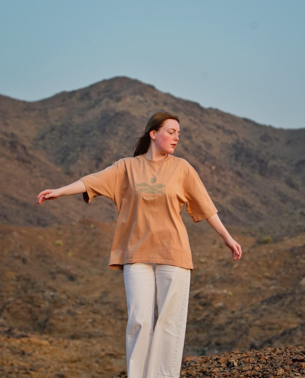 The Empty Quarter tee worn in the mountains, arms out — the tonal embroidery across the chest. Female model wears M in the Oversized fit.