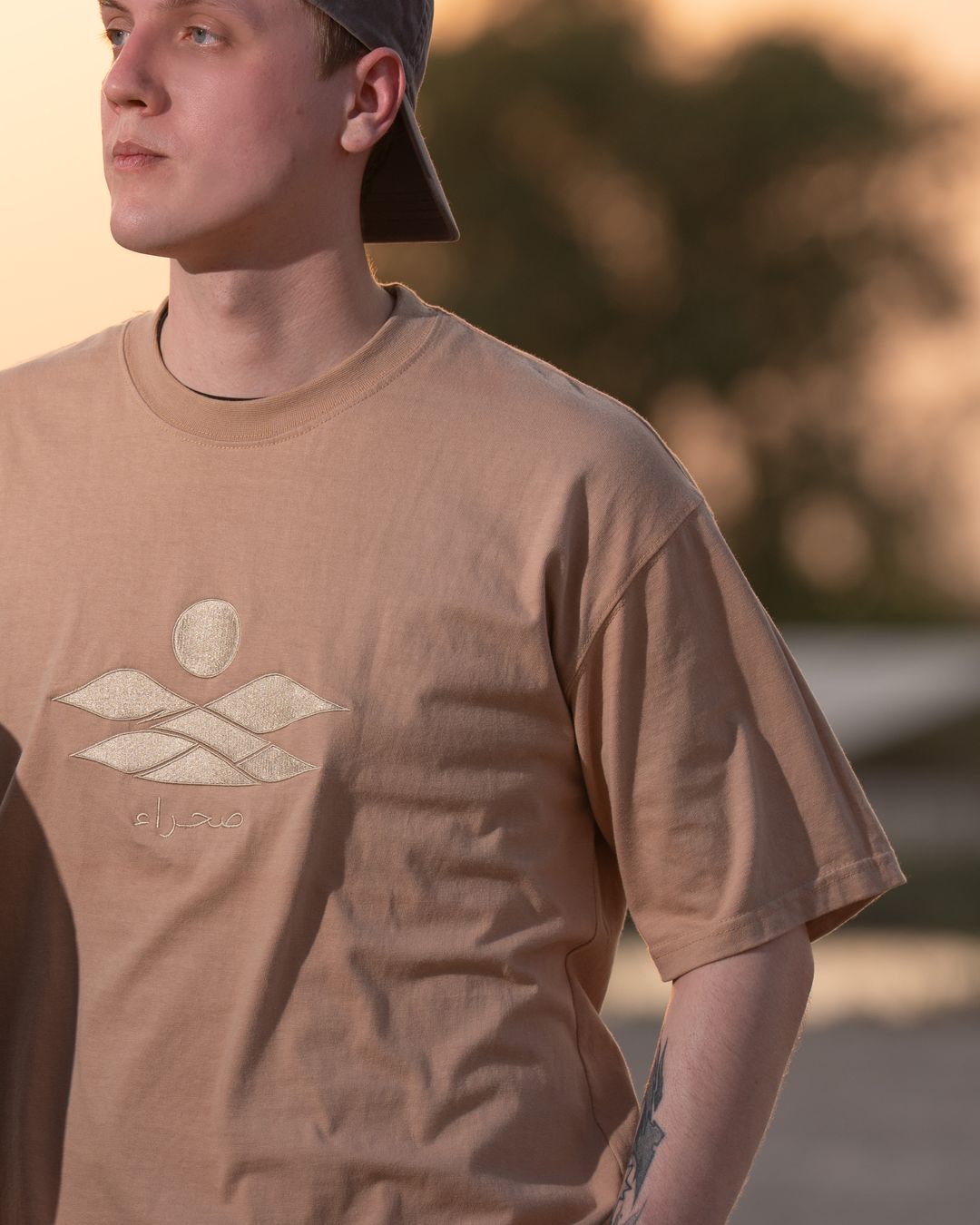 The Empty Quarter tee, front — the tonal embroidered dunes, sun and صحراء on the chest, close. Male model wears M in the Oversized fit.
