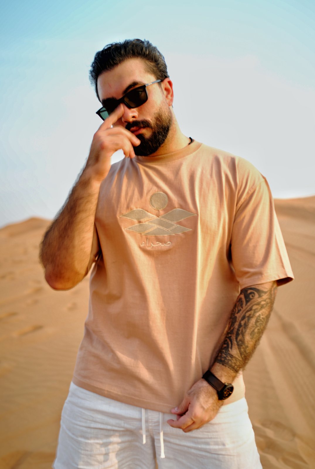 The Empty Quarter tee worn in the dunes — the tonal embroidered dunes, sun and صحراء wordmark across the chest
