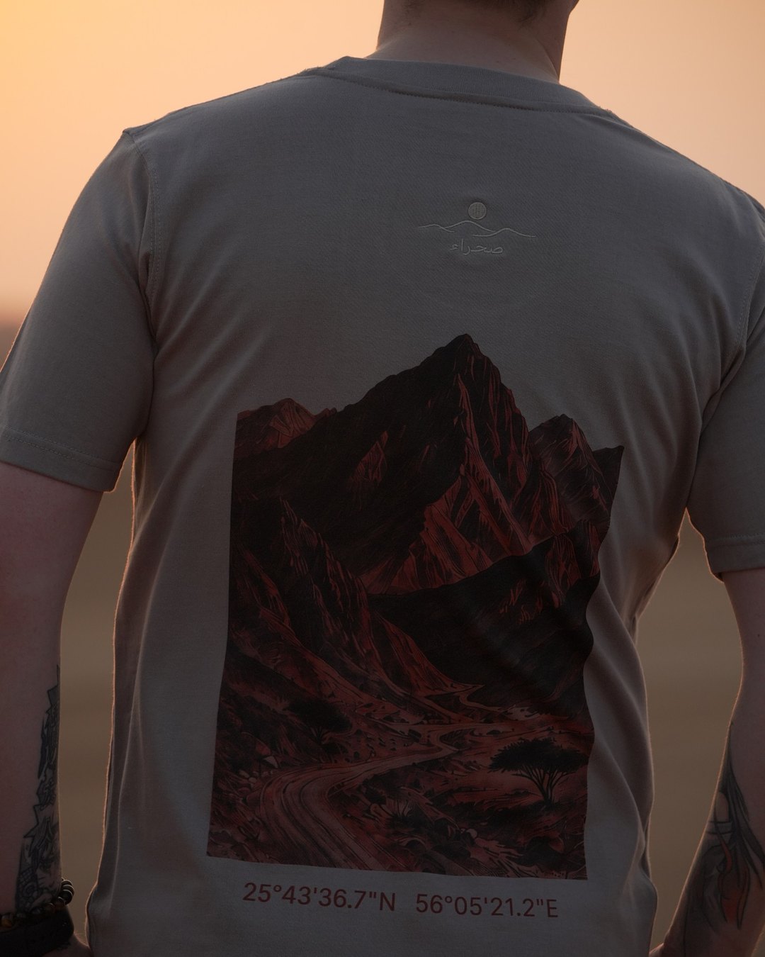 The Hajar Mountains tee worn in the dunes — the printed ridgeline across the back with the coordinates beneath it
