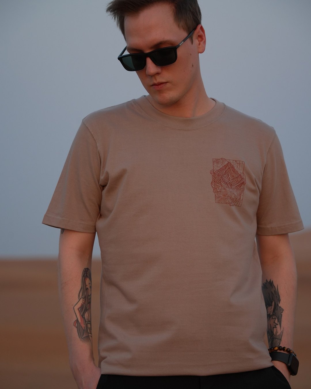 The Hajar Mountains tee, front, worn in the dunes — the chest mark on the sand-coloured cotton