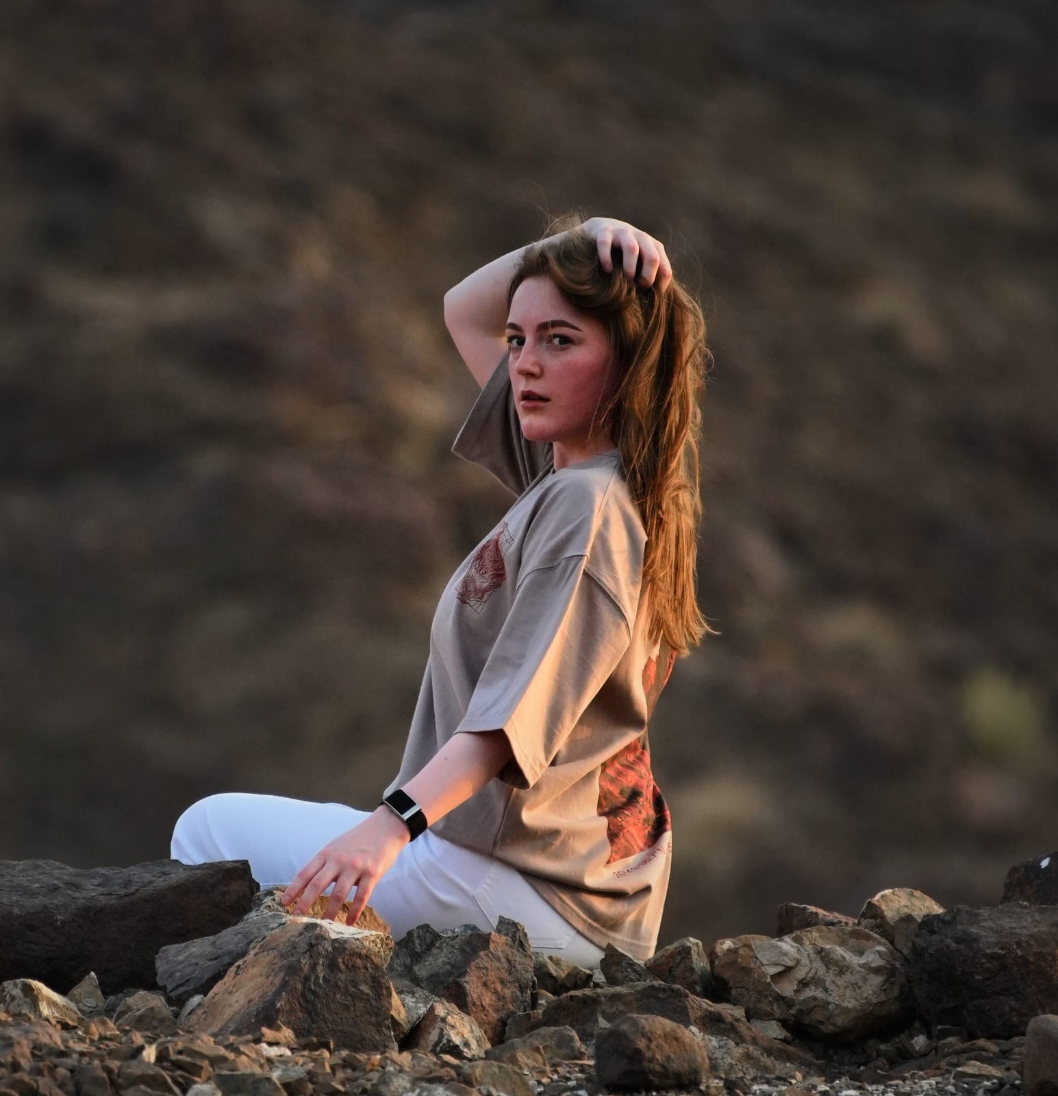 The Hajar Mountains tee worn seated on the rocks at last light — the red-rock print showing at the side. Female model wears M in the Oversized fit.