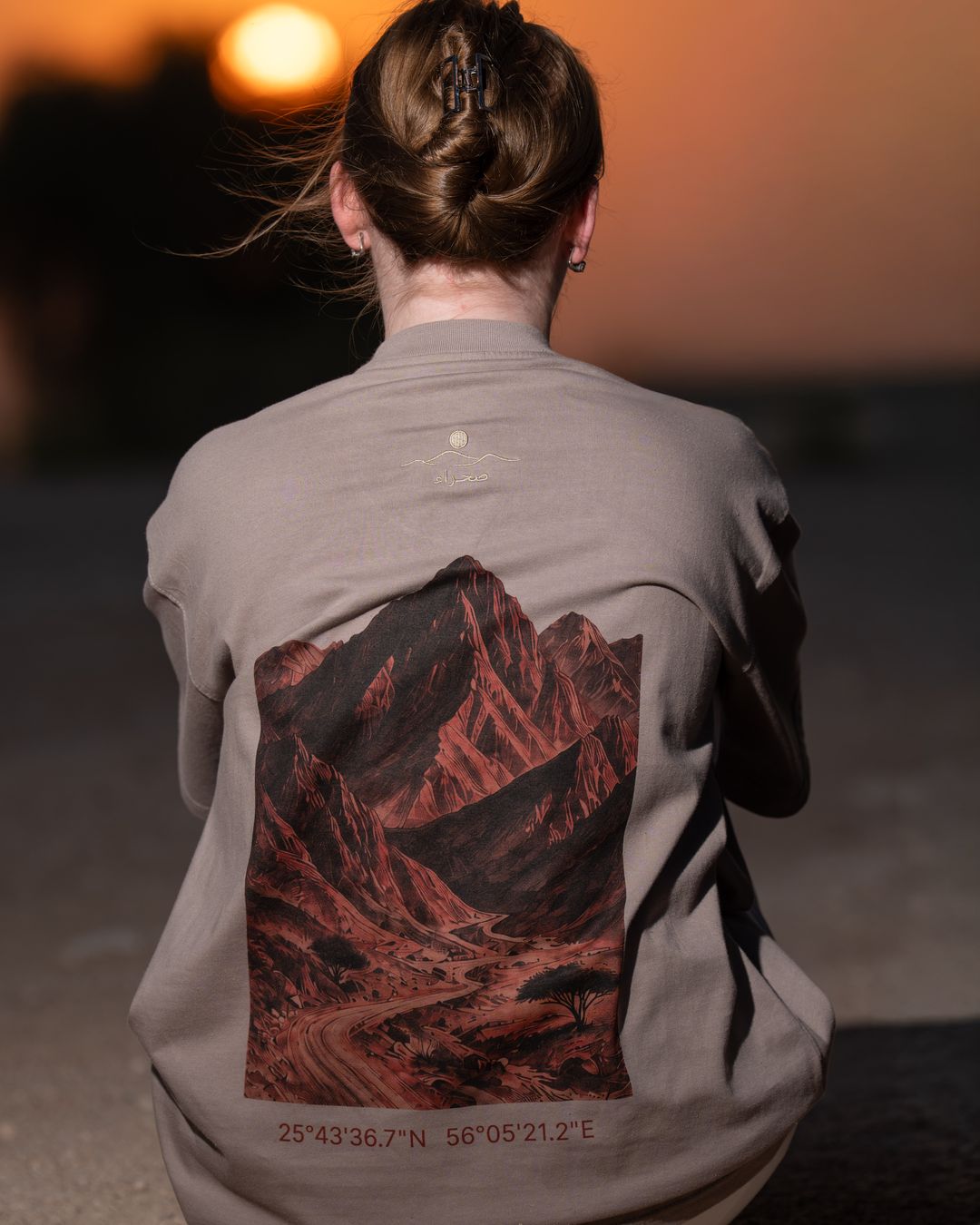 The Hajar Mountains tee from behind at sunset — the mountain print and coordinates filling the back. Female model wears M in the Oversized fit.