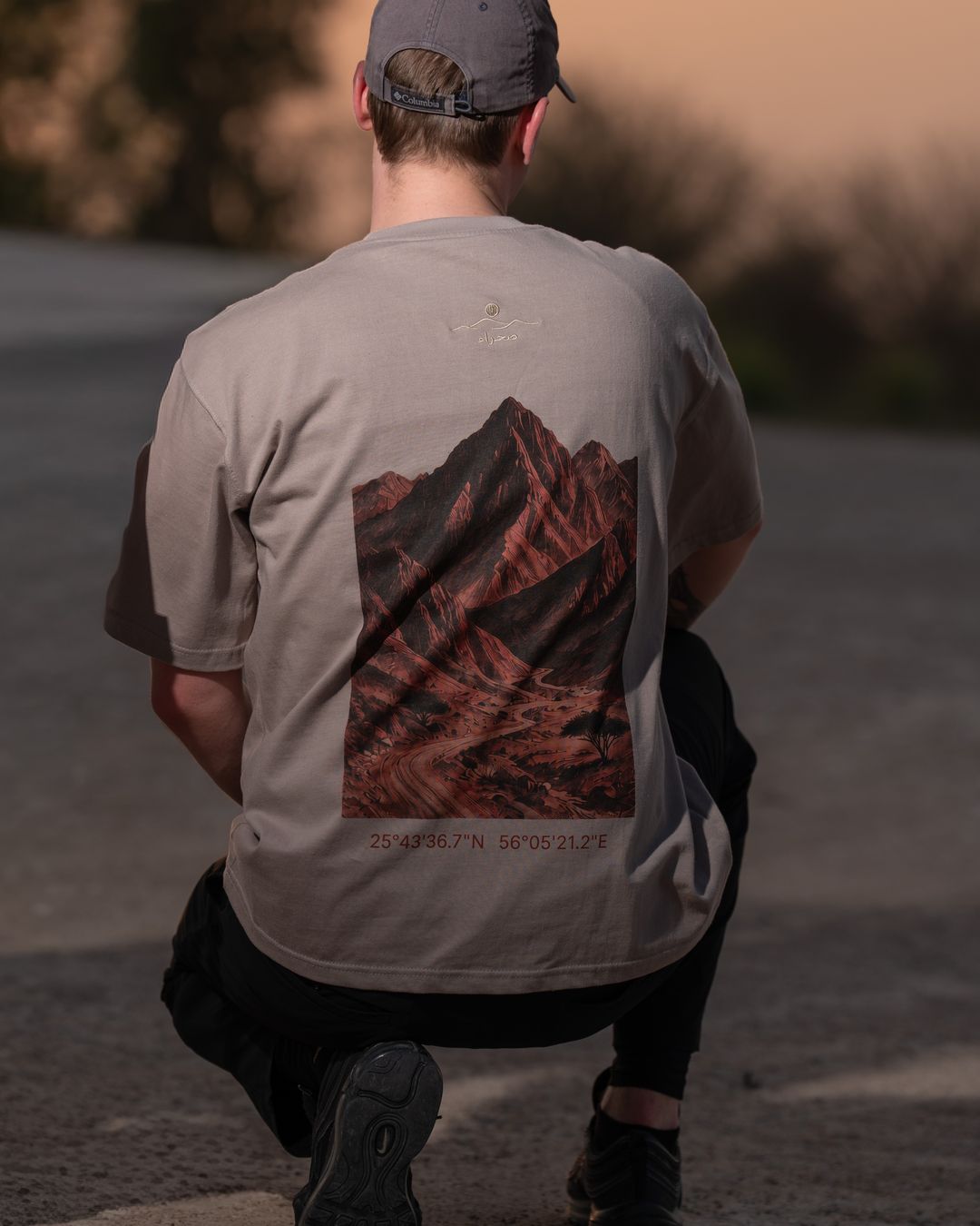 The Hajar Mountains tee from behind, kneeling — the mountain print and the coordinates beneath it. Male model wears M in the Oversized fit.