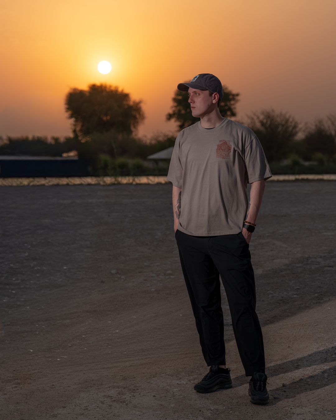 The Hajar Mountains tee worn full-length with the sun behind the trees — the chest mark on the front. Male model wears M in the Oversized fit.