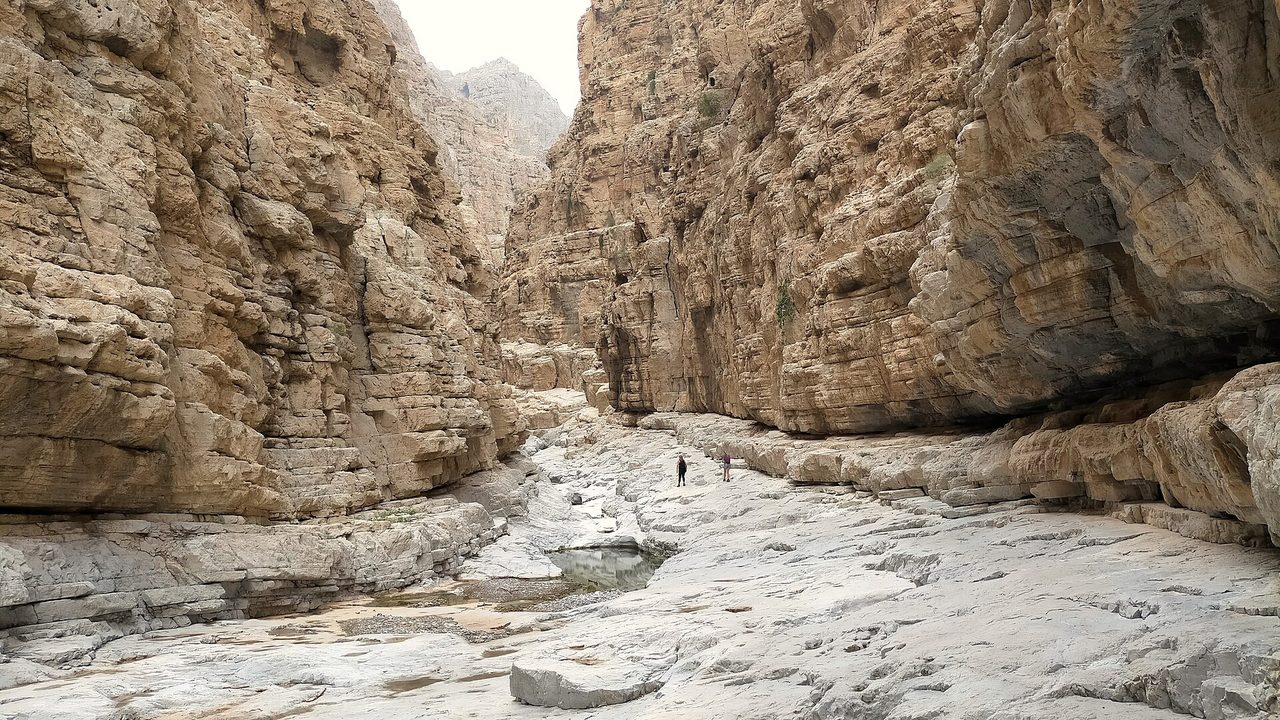 Two small figures walking along a pale rock floor between tall limestone walls in a canyon in Wadi Naqab