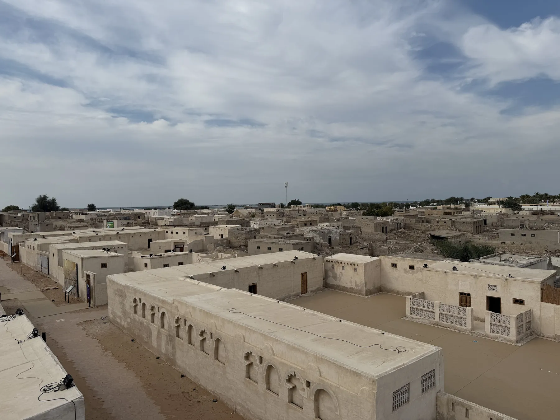 Flat-roofed coral-stone and plaster buildings of Al Jazirah Al Hamra seen from above, with a cloudy sky over the rooftops